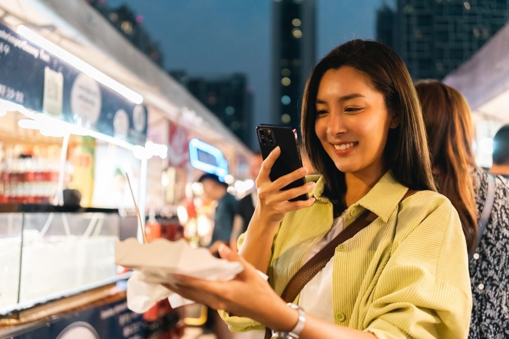 หญิงสาวถ่ายรูปสตรีตฟูดที่ซื้อจากตลาดนัดกลางคืนกรุงเทพฯ